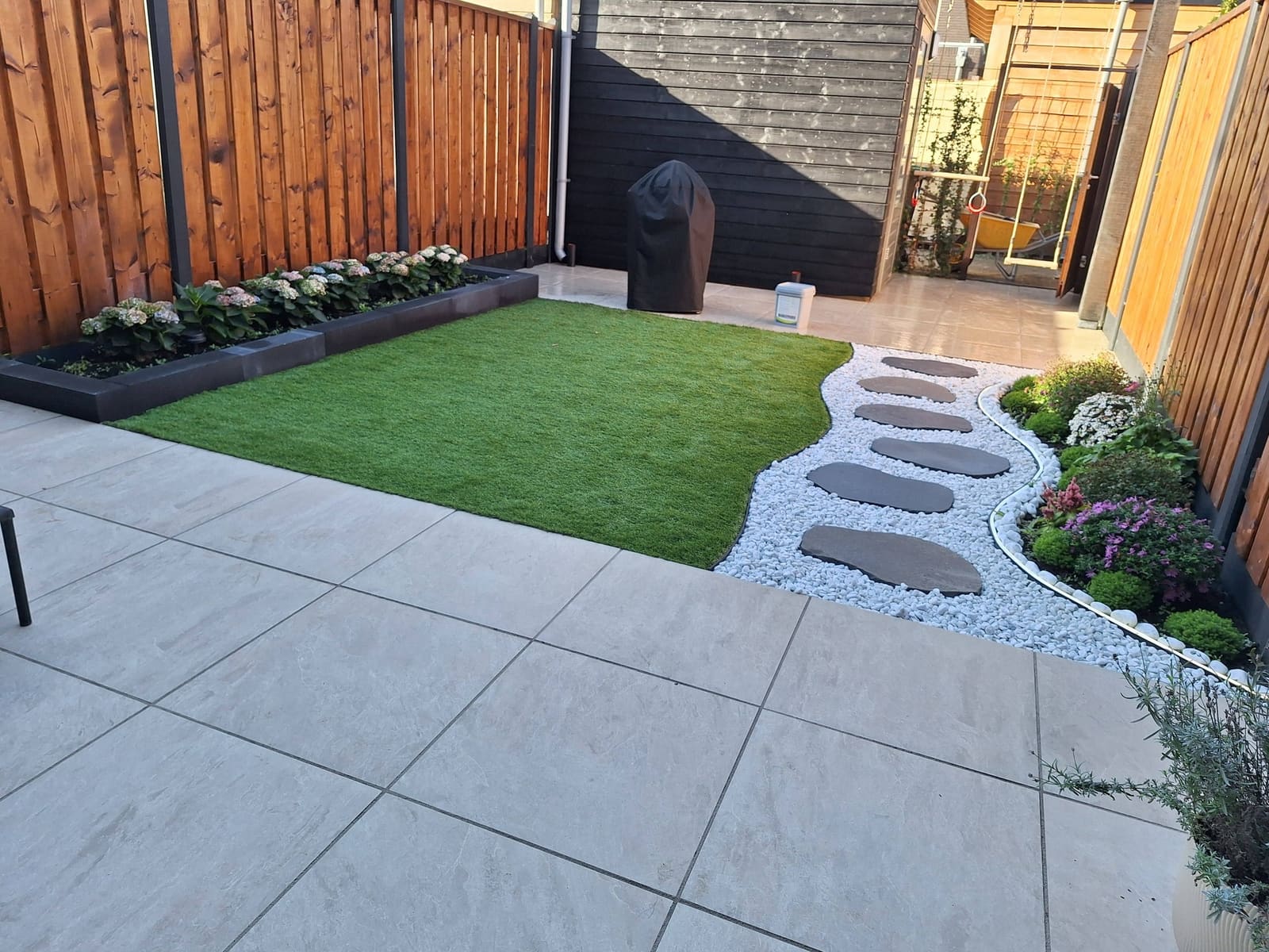 Achtertuin met kunstgras, een rand van wit grind met stapstenen en een terras