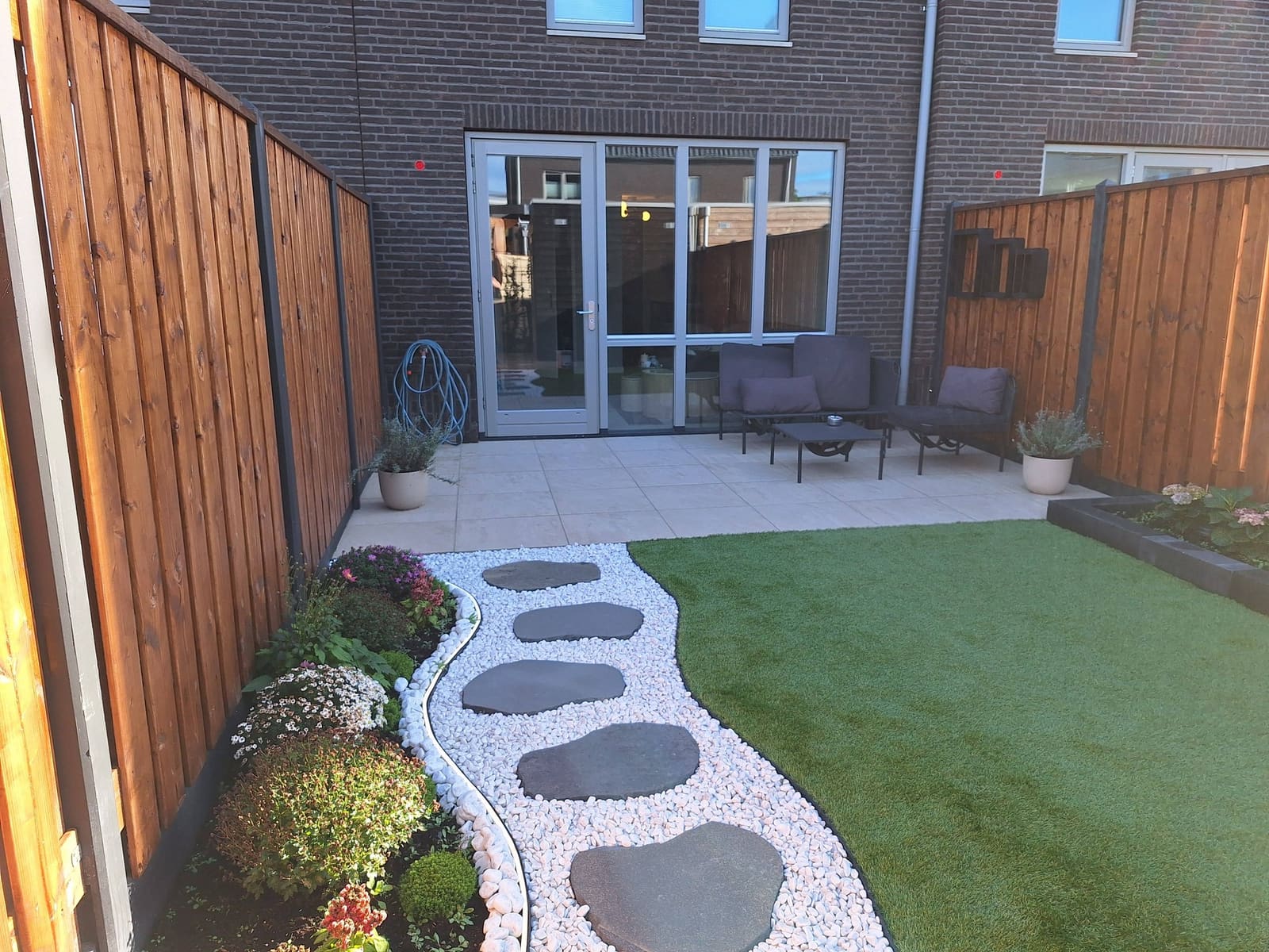 Pad van donkere stapstenen in wit grind naar een terras met loungeset, met kunstgras en een border langs de houten schutting