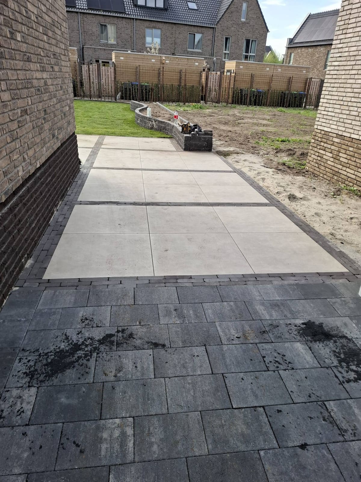 Tegelpad langs een nieuwbouwwoning tijdens de aanleg, met zand en gereedschap