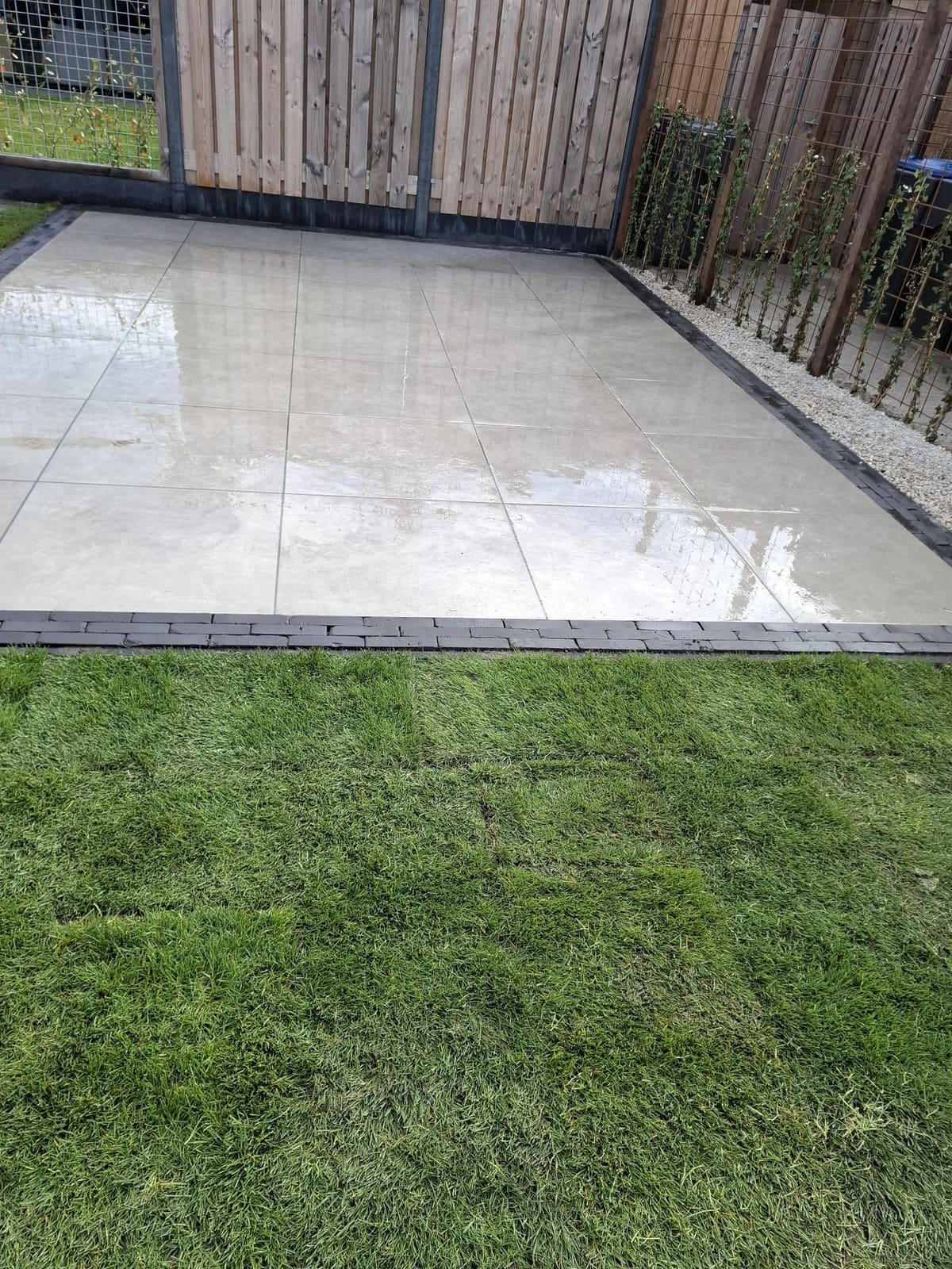 Nieuw terras van grote glanzende tegels met een rand, ernaast vers gras, langs een houten schutting