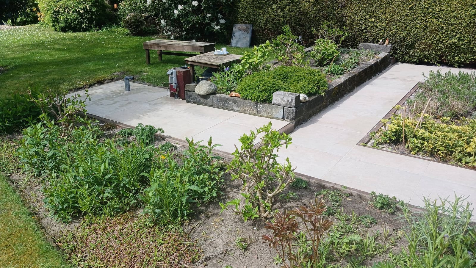 Tuin met paden van lichte tegels rond borders met nieuwe beplanting en een zitplek